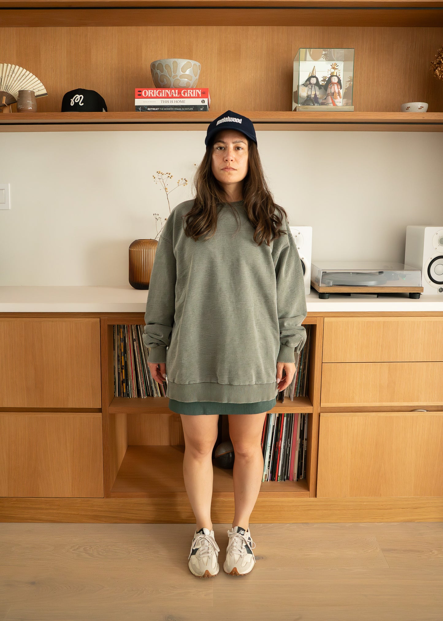 Woman wearing an oversized green sweatshirt and skirt standing in front of a wooden shelf.