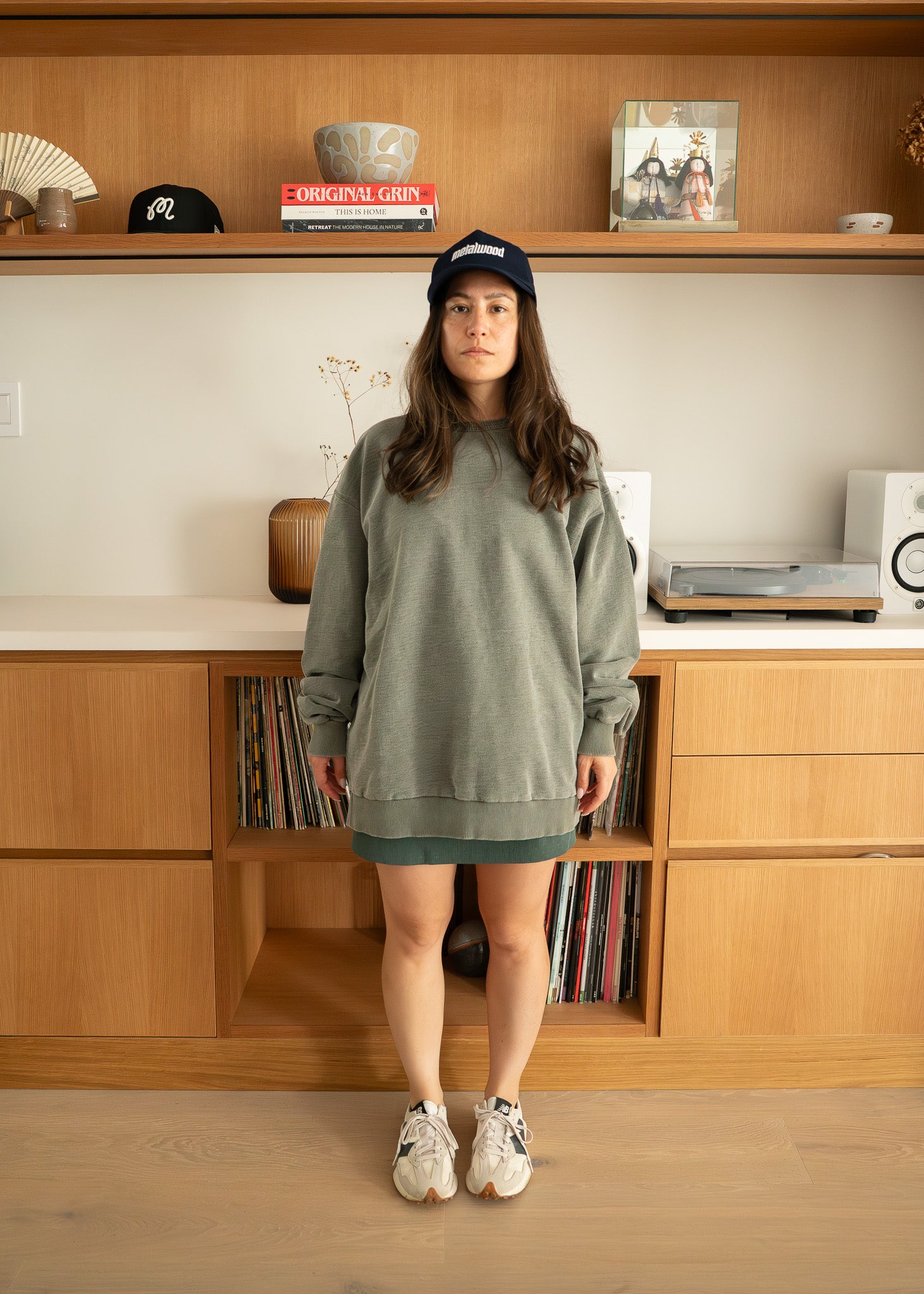 Woman wearing an oversized green sweatshirt and skirt standing in front of a wooden shelf.