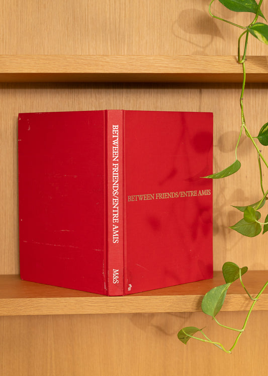 Vintage red book titled 'Between Friends/Entre Amis' on a wooden shelf with a plant.
