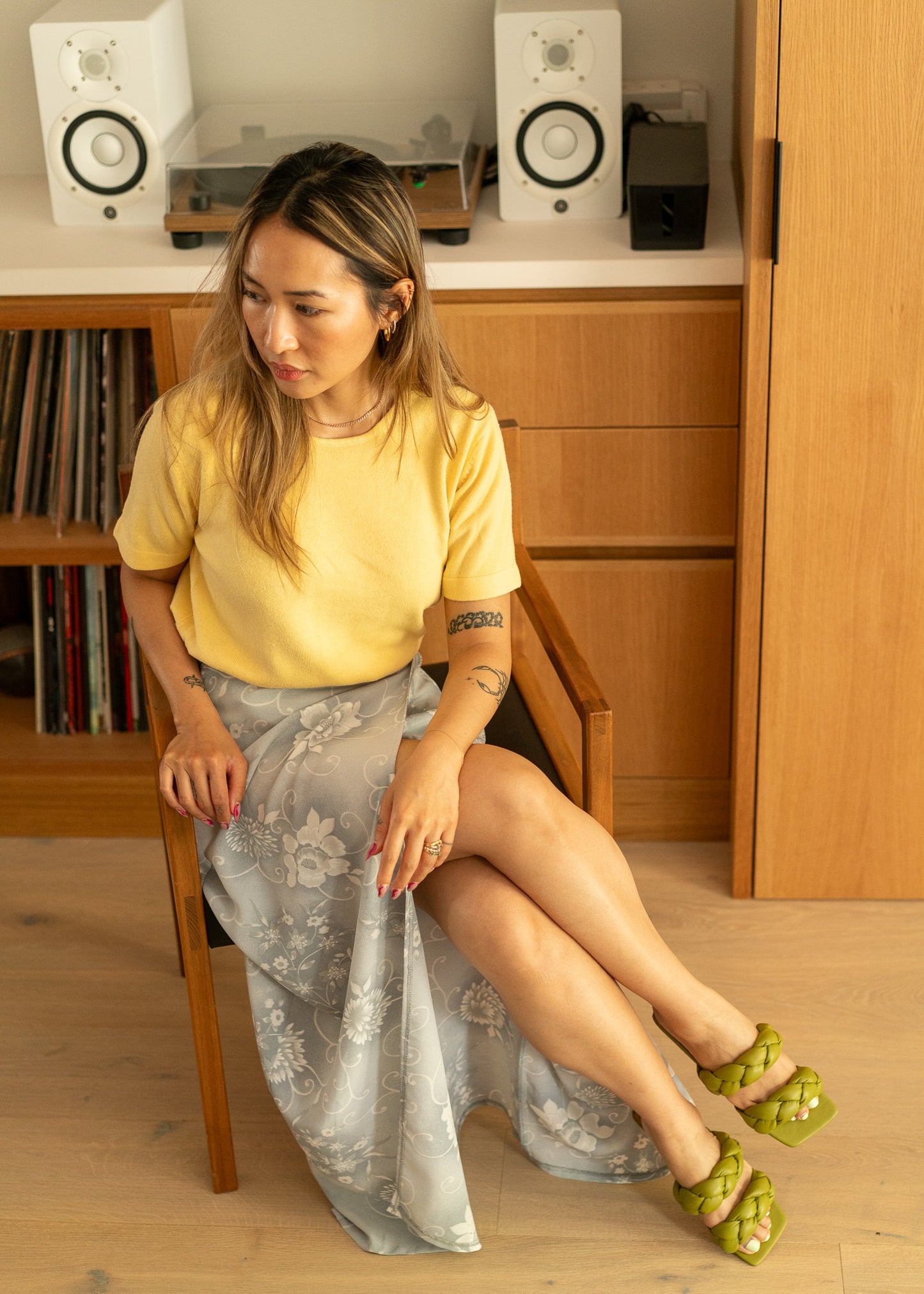 Woman wearing a vintage yellow short sleeve sweater and light grey floral skirt sitting on a chair in a room with wooden furniture and speakers.