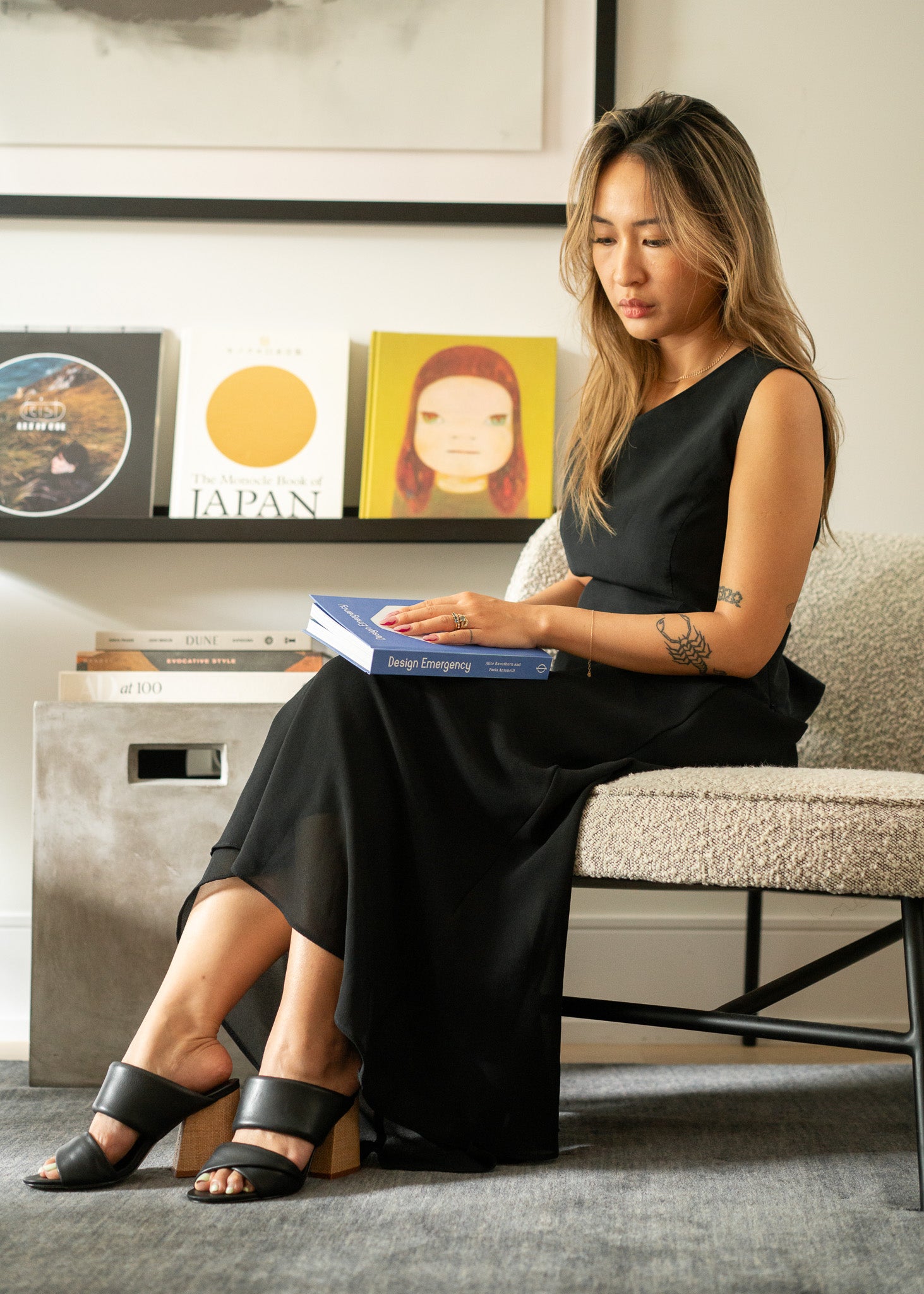 Woman wearing a sleeveless black top and long black skirt sitting on a chair reading a book in a room with books and decor on a shelf.