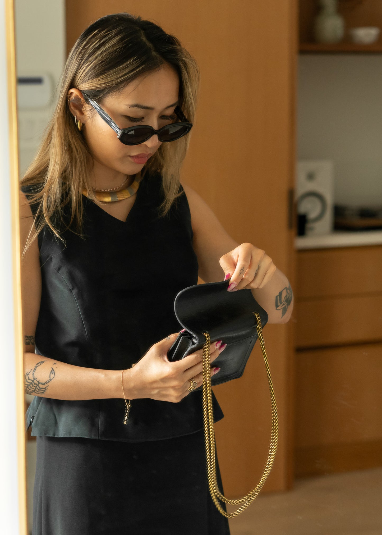 Woman wearing a fitted black tank top holding a black handbag with gold chain strap indoors.
