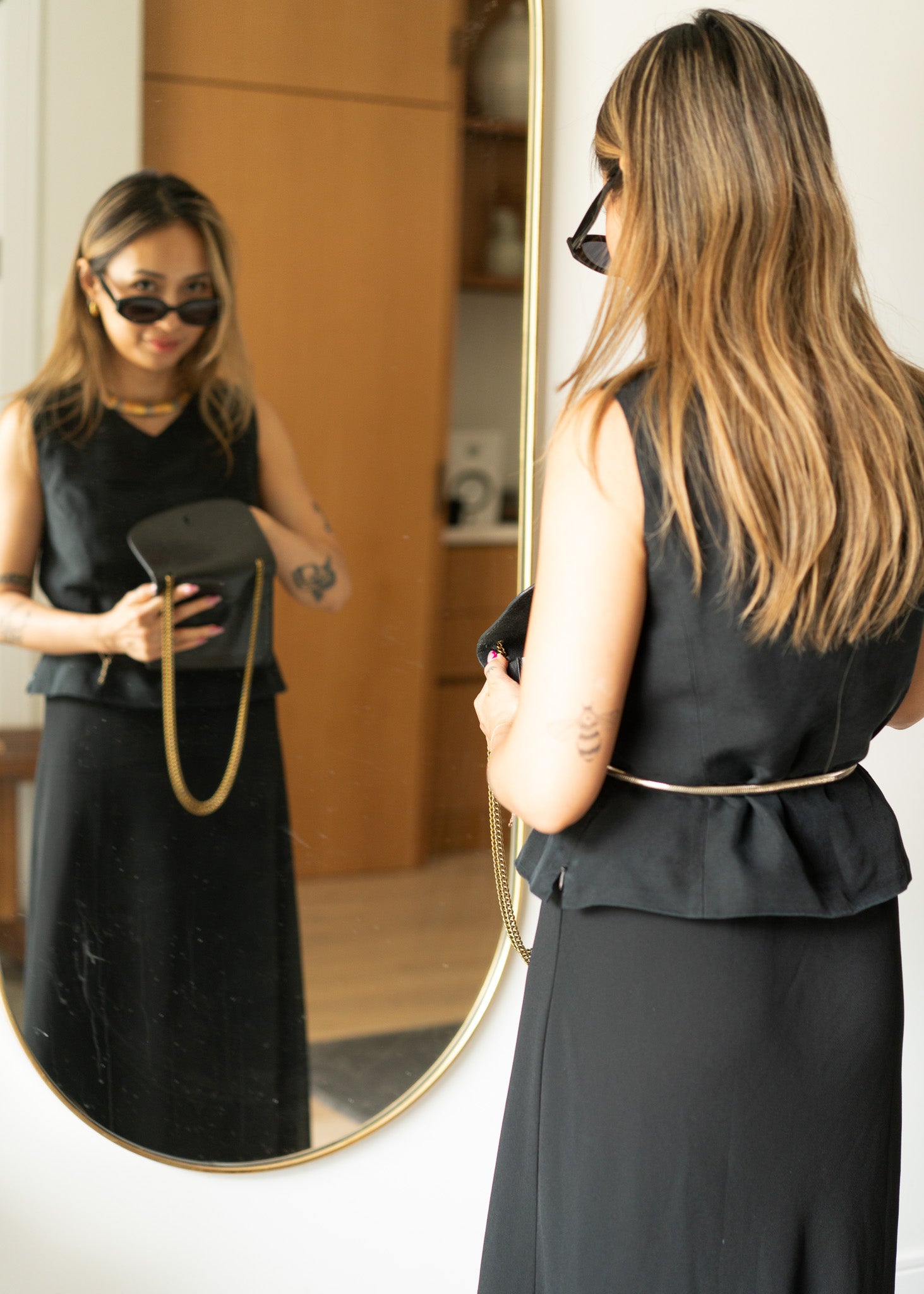 Woman in a black sleeveless top and long black skirt looking at herself in a mirror.