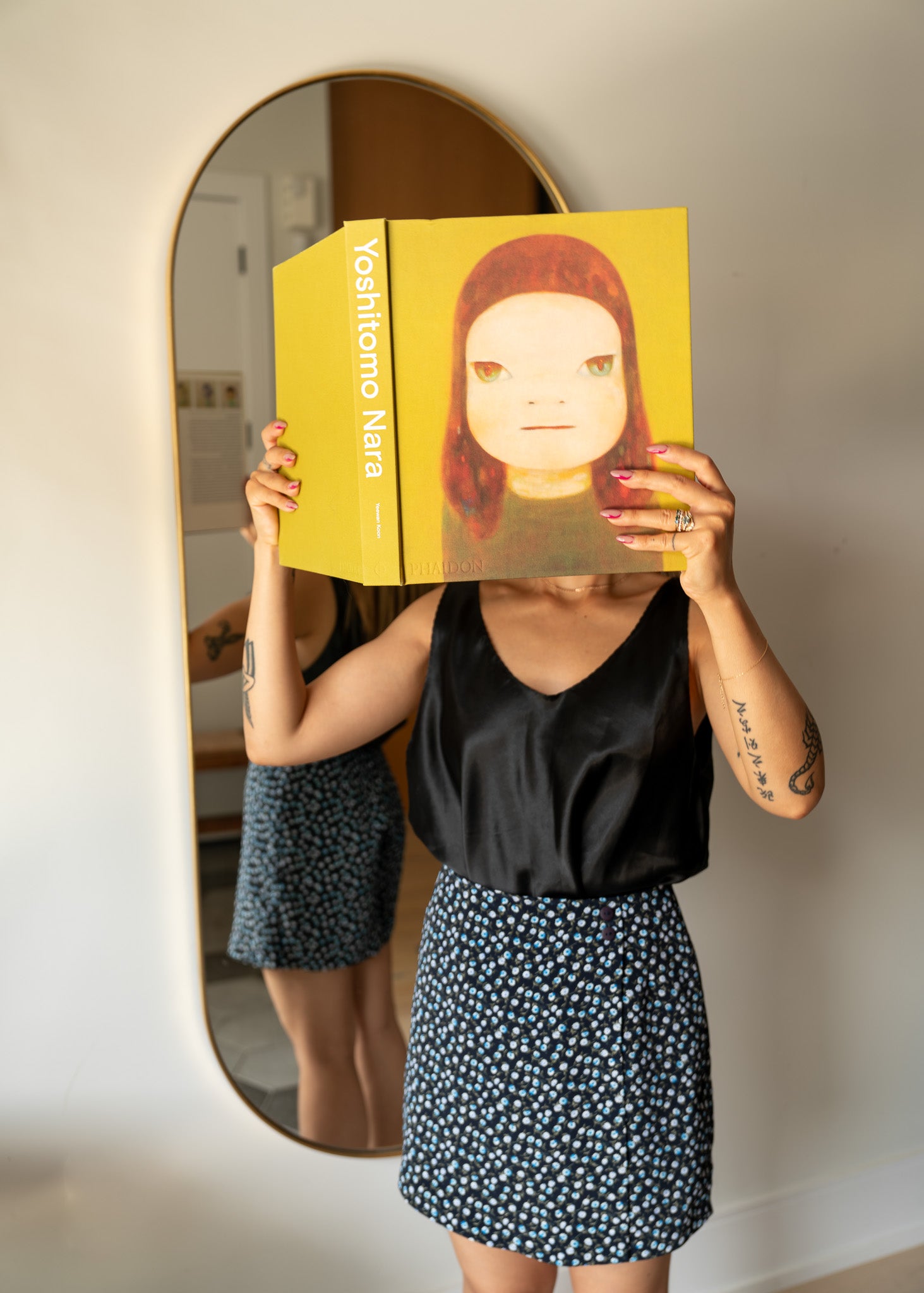 Person holding a book with a yellow cover featuring a woman's face in front of their face, standing in front of a mirror.
