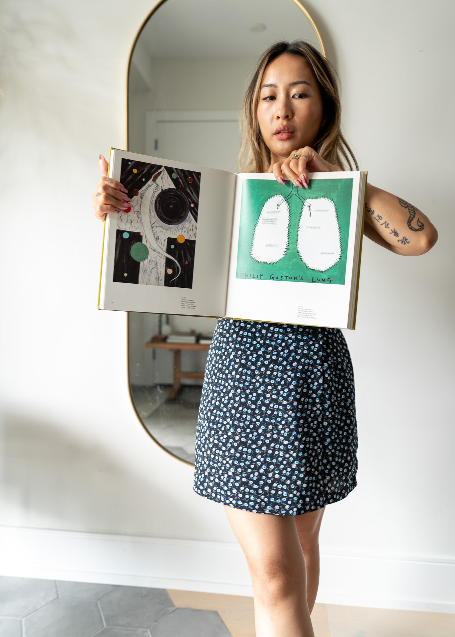 Person wearing a blue floral miniskirt holding an art book in front of a mirror