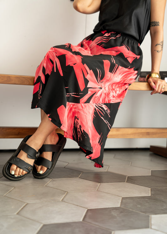 Person wearing a black and pink floral skirt and brass bangle sitting on a wooden bench.