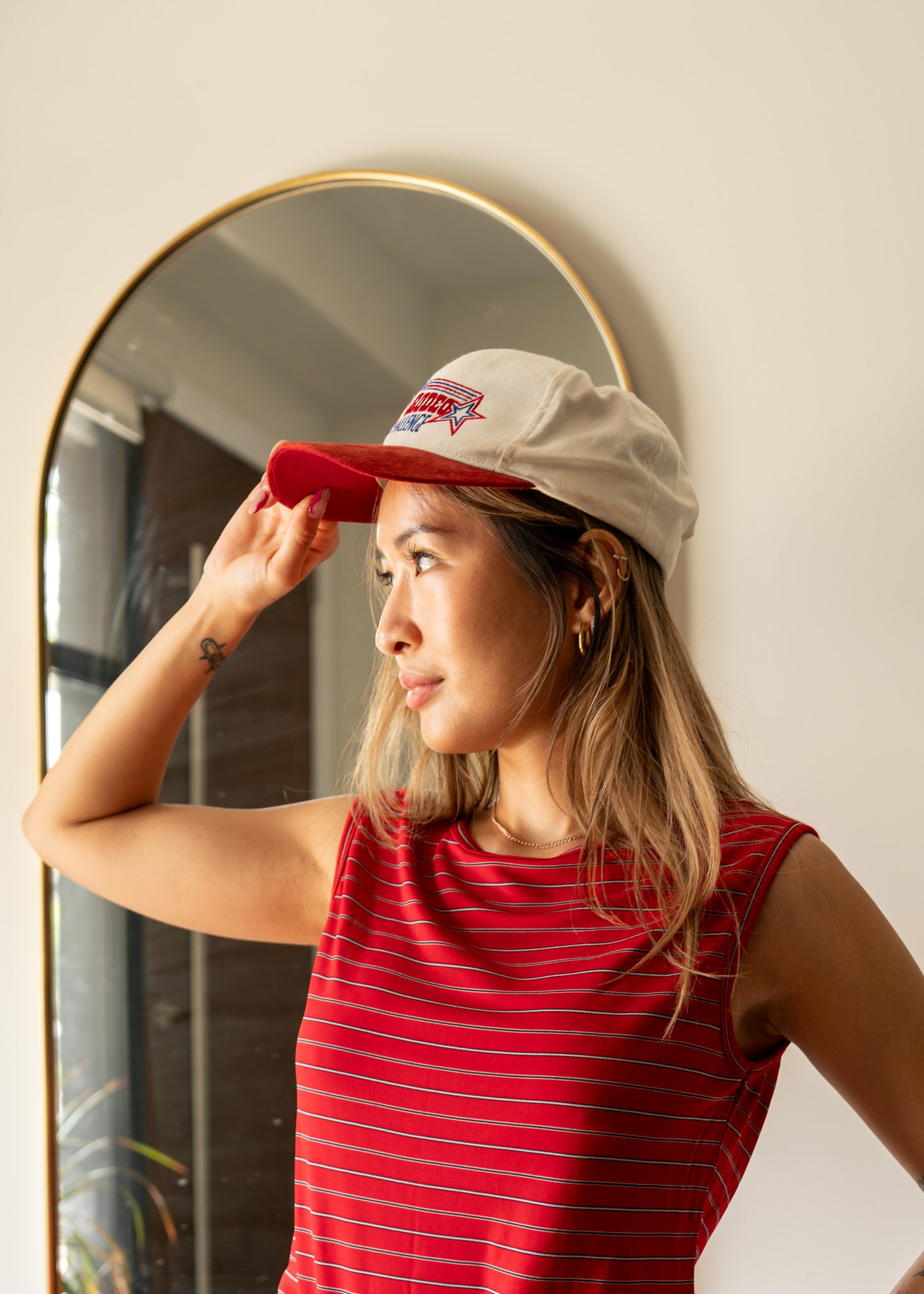 Woman adjusting a red suede cap in front of a mirror, wearing a red striped tank top.