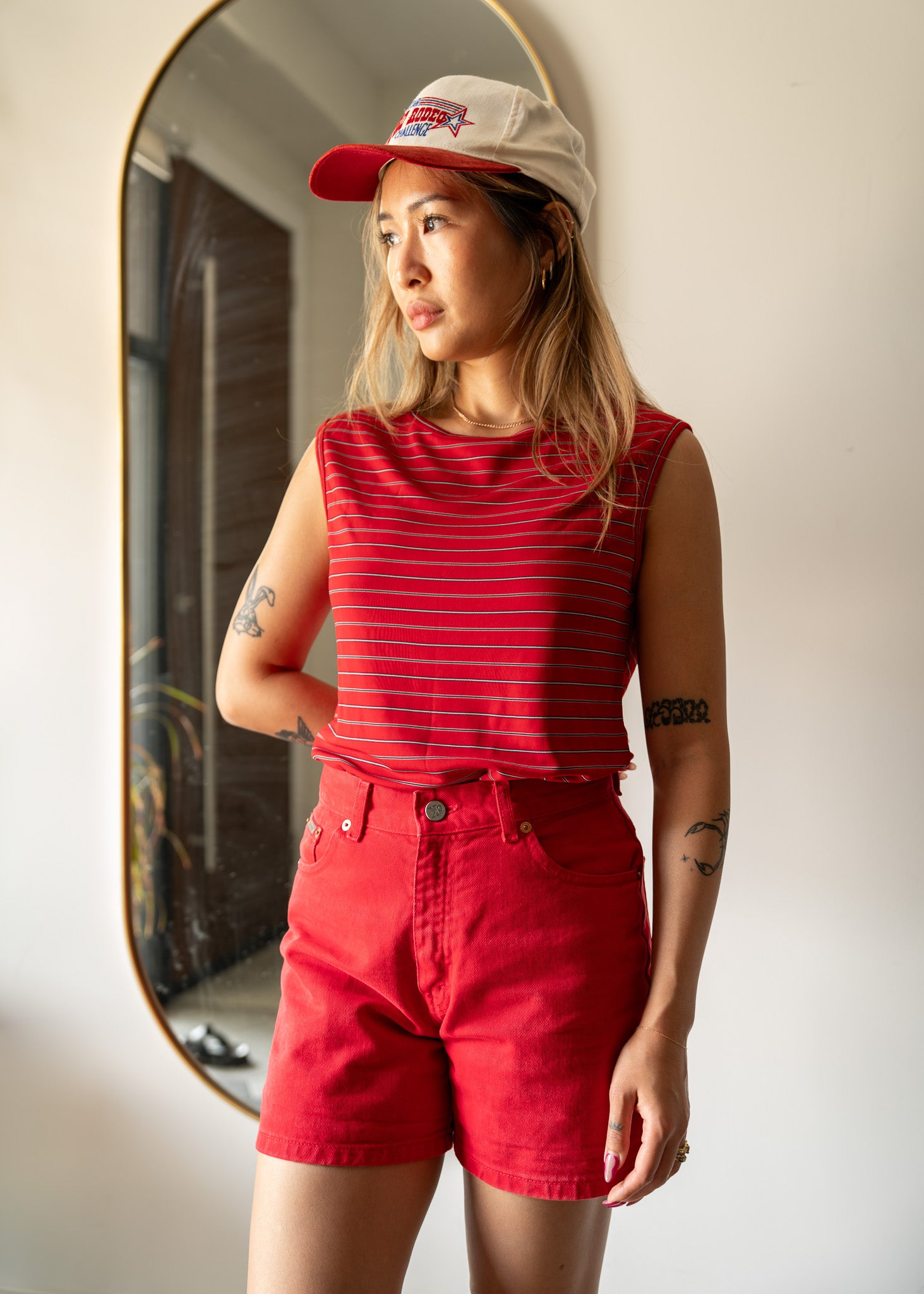 Person wearing a red striped tank top and shorts with a red suede and cotton cap, standing in front of a mirror.