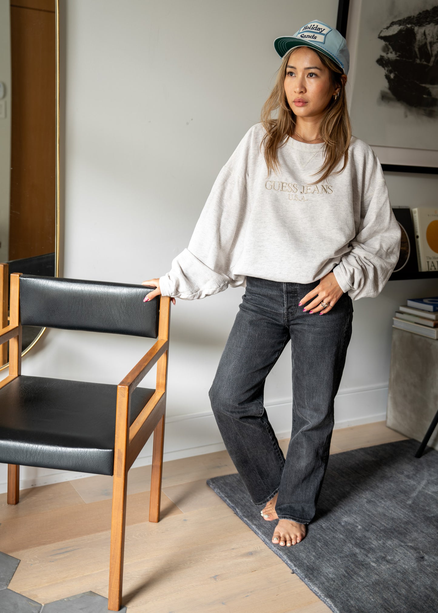Person wearing a light grey sweatshirt and light blue Holiday Sands cap beside a leather and wooden chair
