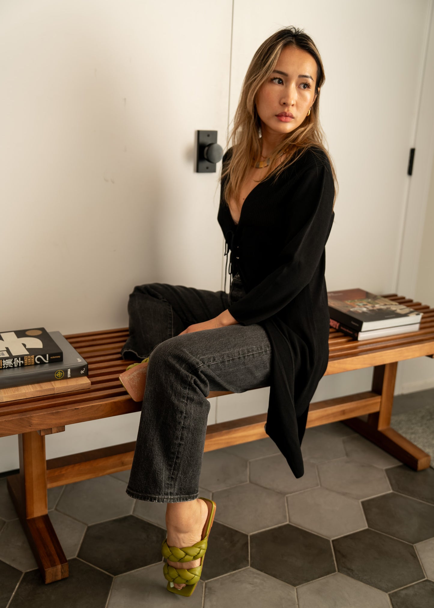 Woman sitting on a wooden bench with books around her, wearing a black tie front cardigan and black jeans