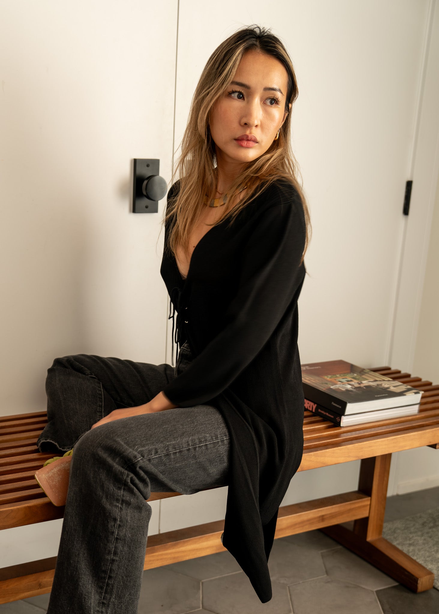 Woman sitting on a wooden bench wearing a black cardigan and black jeans with a white wall background
