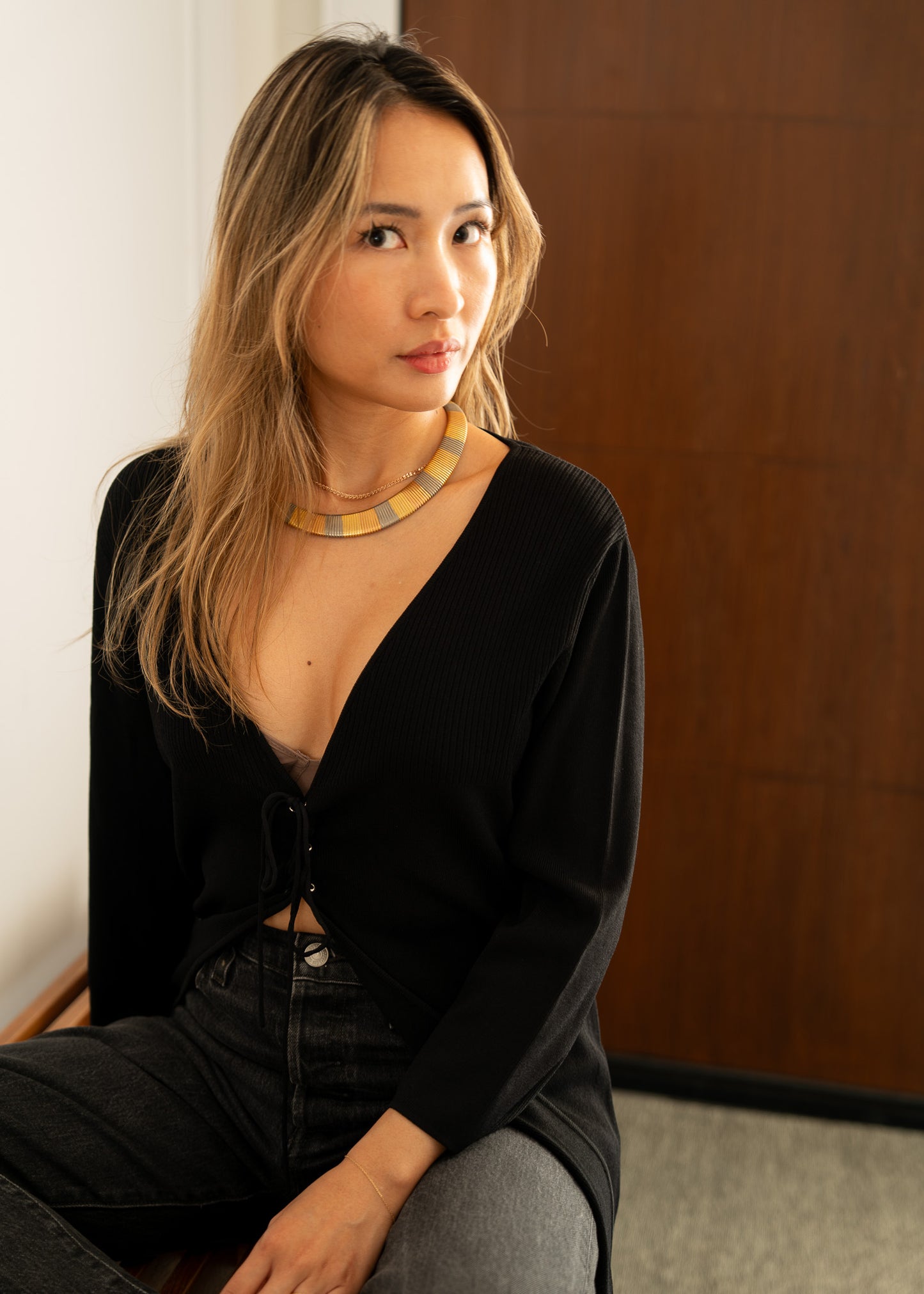Woman wearing a black outfit with a deep v neckline and metal necklace, sitting against a wooden wall.