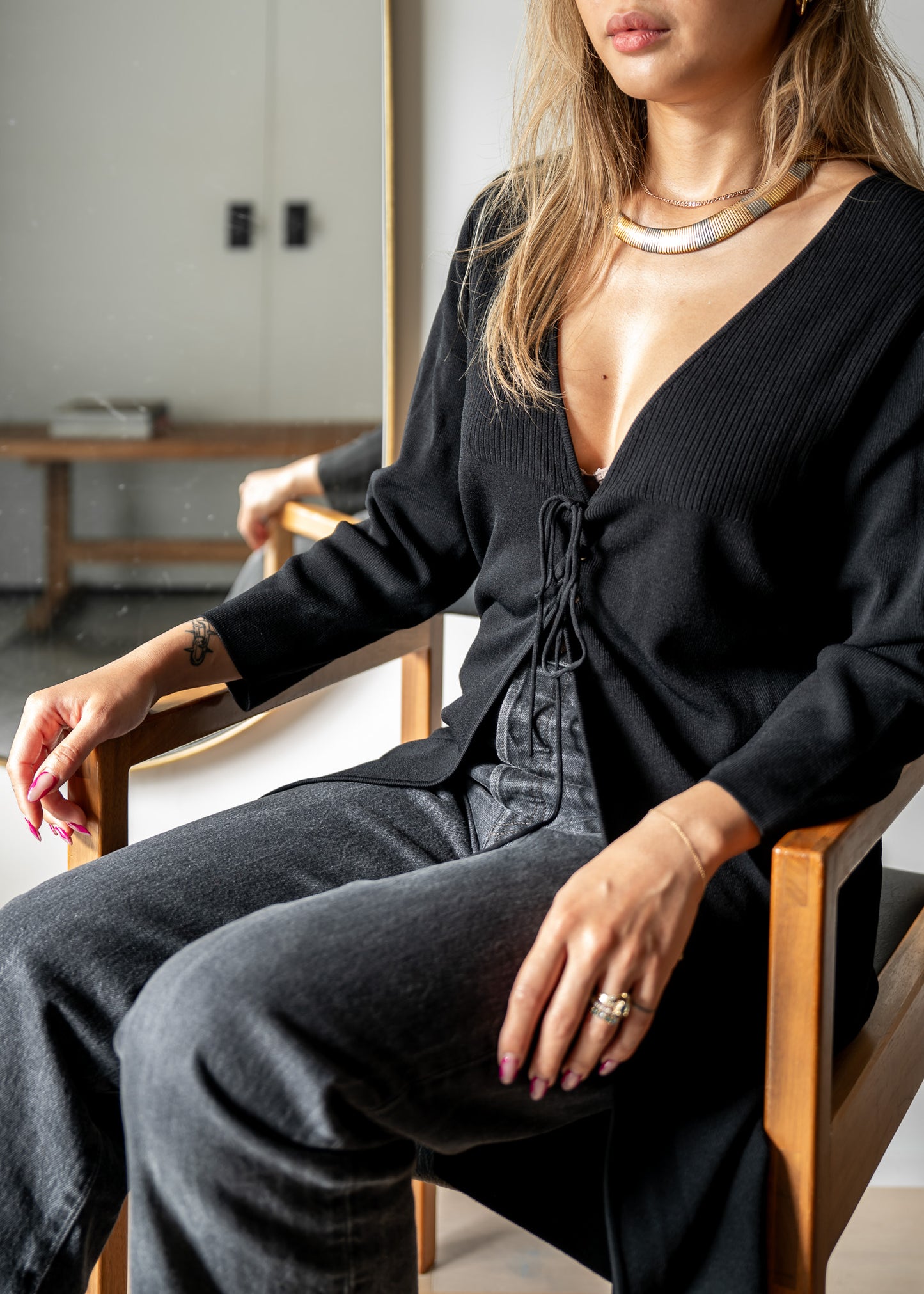 Person wearing a black cardigan and dark jeans sitting on a chair indoors.