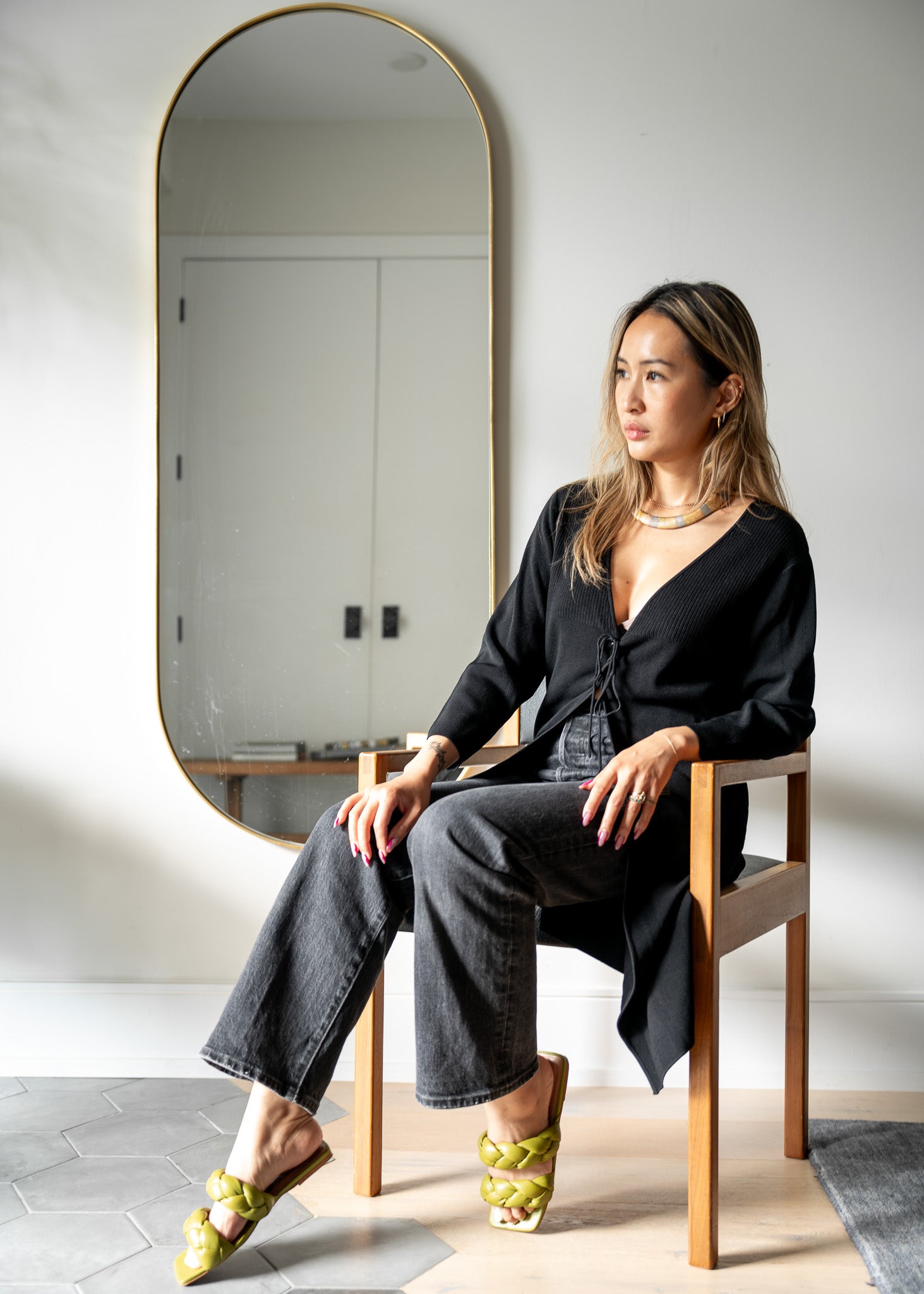 Woman sitting on a chair in front of a tall mirror wearing a black tie front cardigan and black denim jeans with green shoes.
