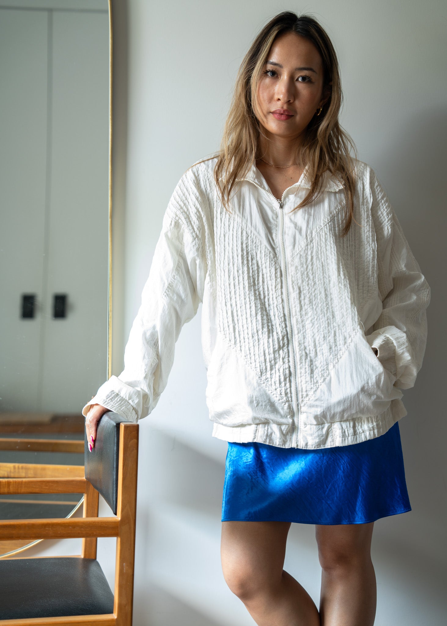Woman wearing a white textured windbreaker and blue skirt standing indoors.