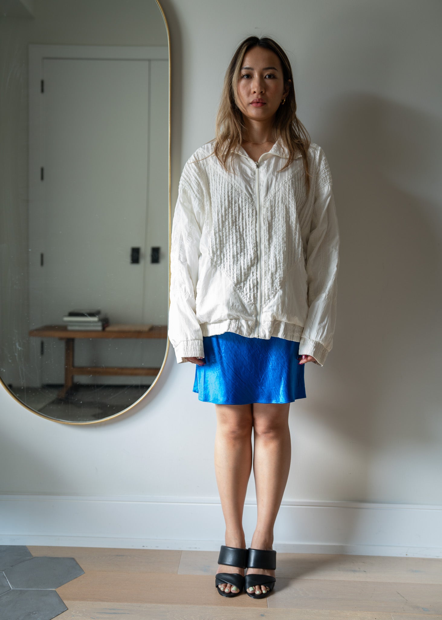 Woman wearing a white textured jacket and blue skirt standing in front of a mirror.
