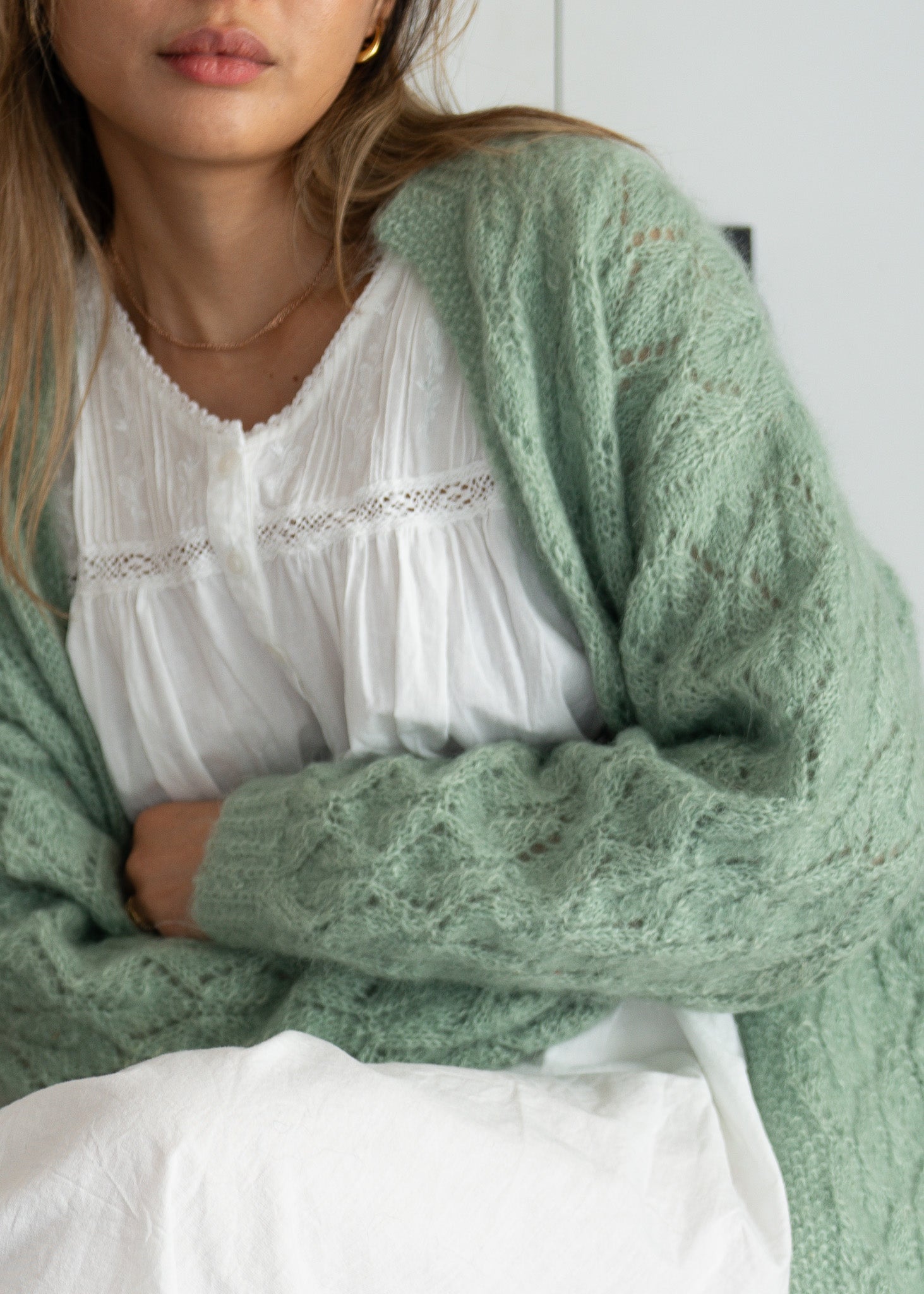 Person wearing a green knitted cardigan over a white cotton dress with a blurred background