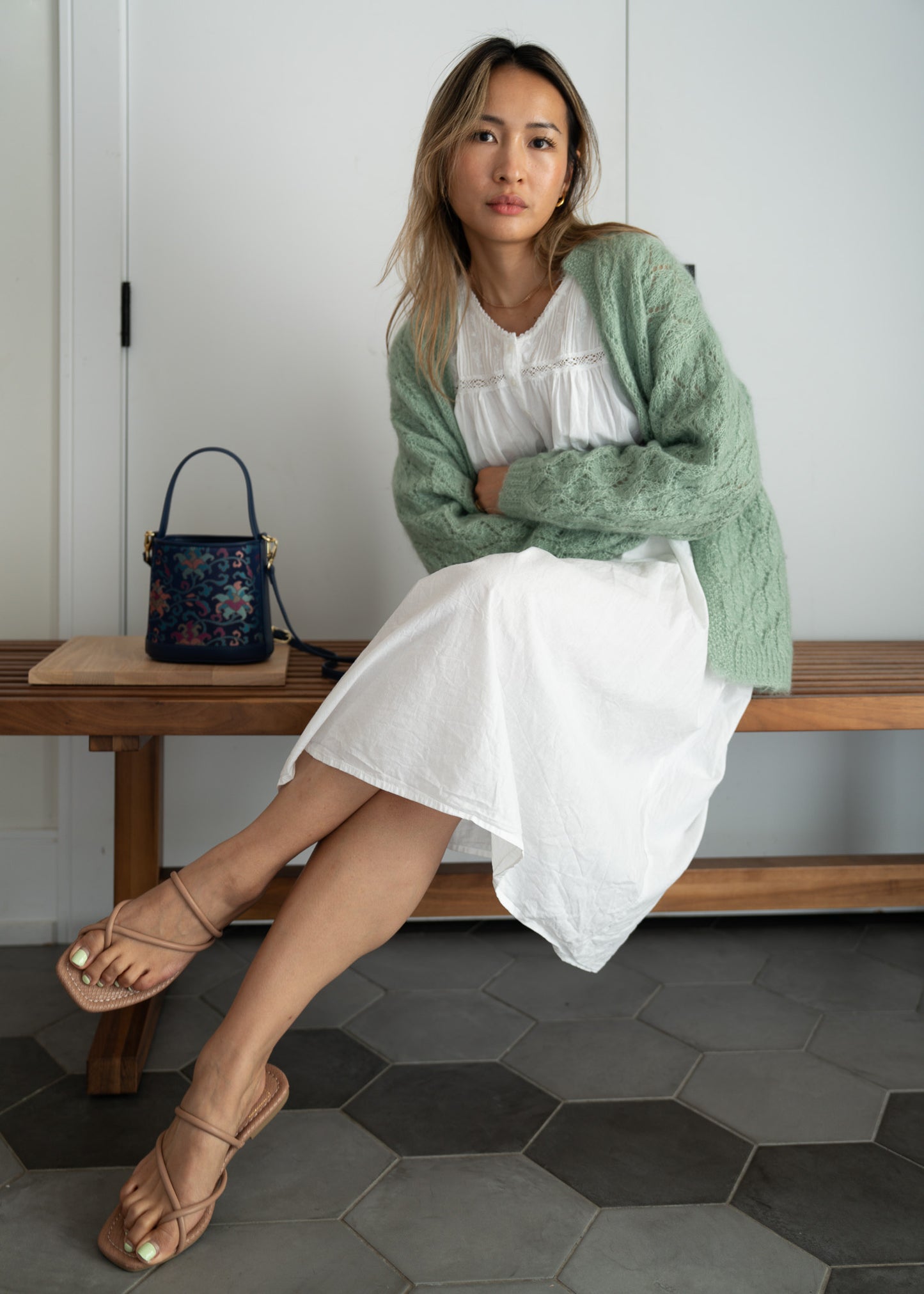 Woman sitting on a wooden bench wearing a green sweater and white dress, with a small bag beside her.