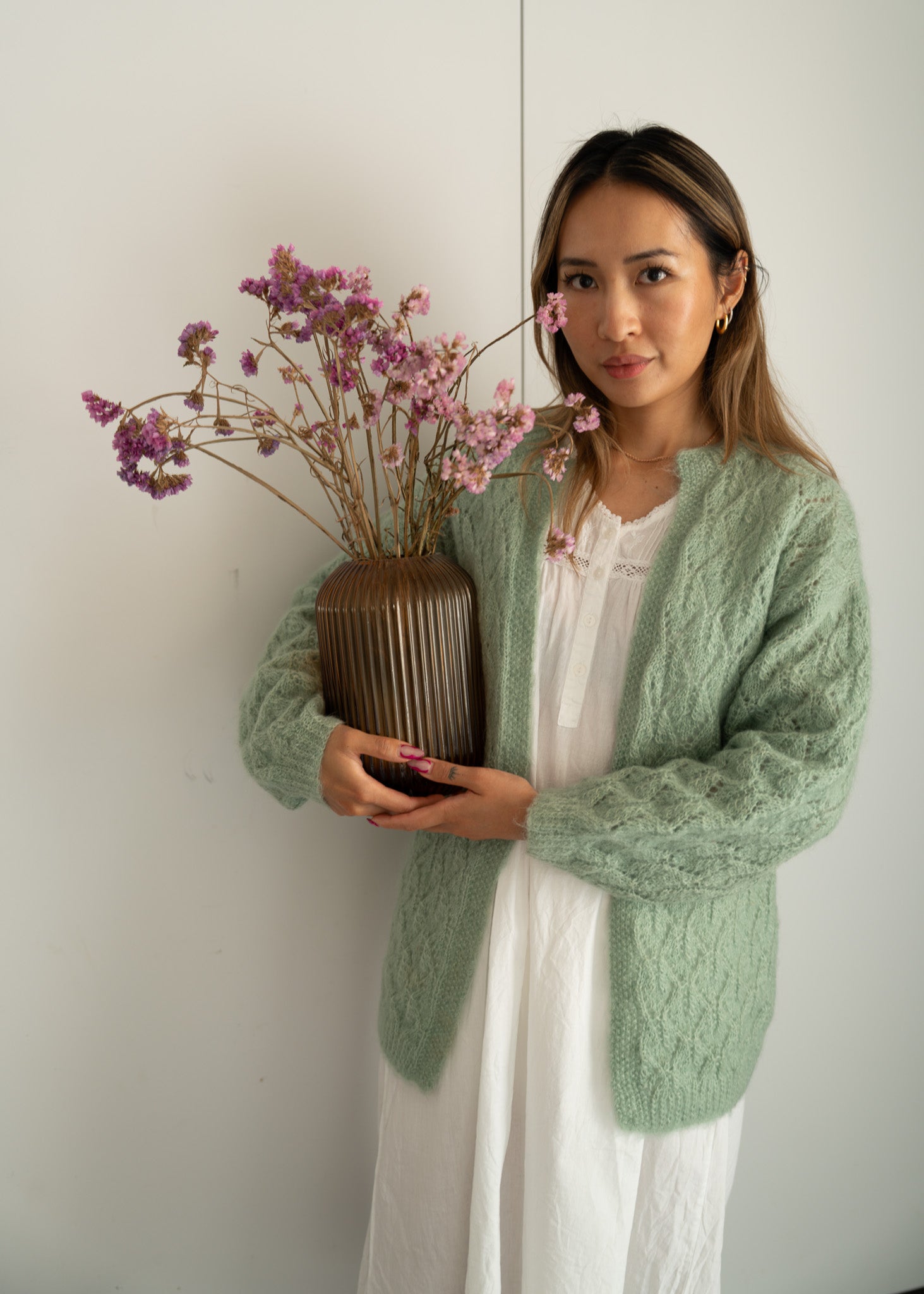 Woman wearing a green cardigan and white dress holding a vase with pink flowers against a white wall