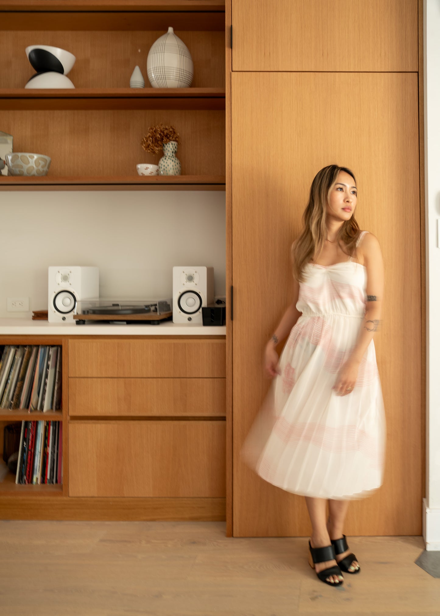 Woman in a white and pink pleated dress standing in a room with wooden shelves and decor.