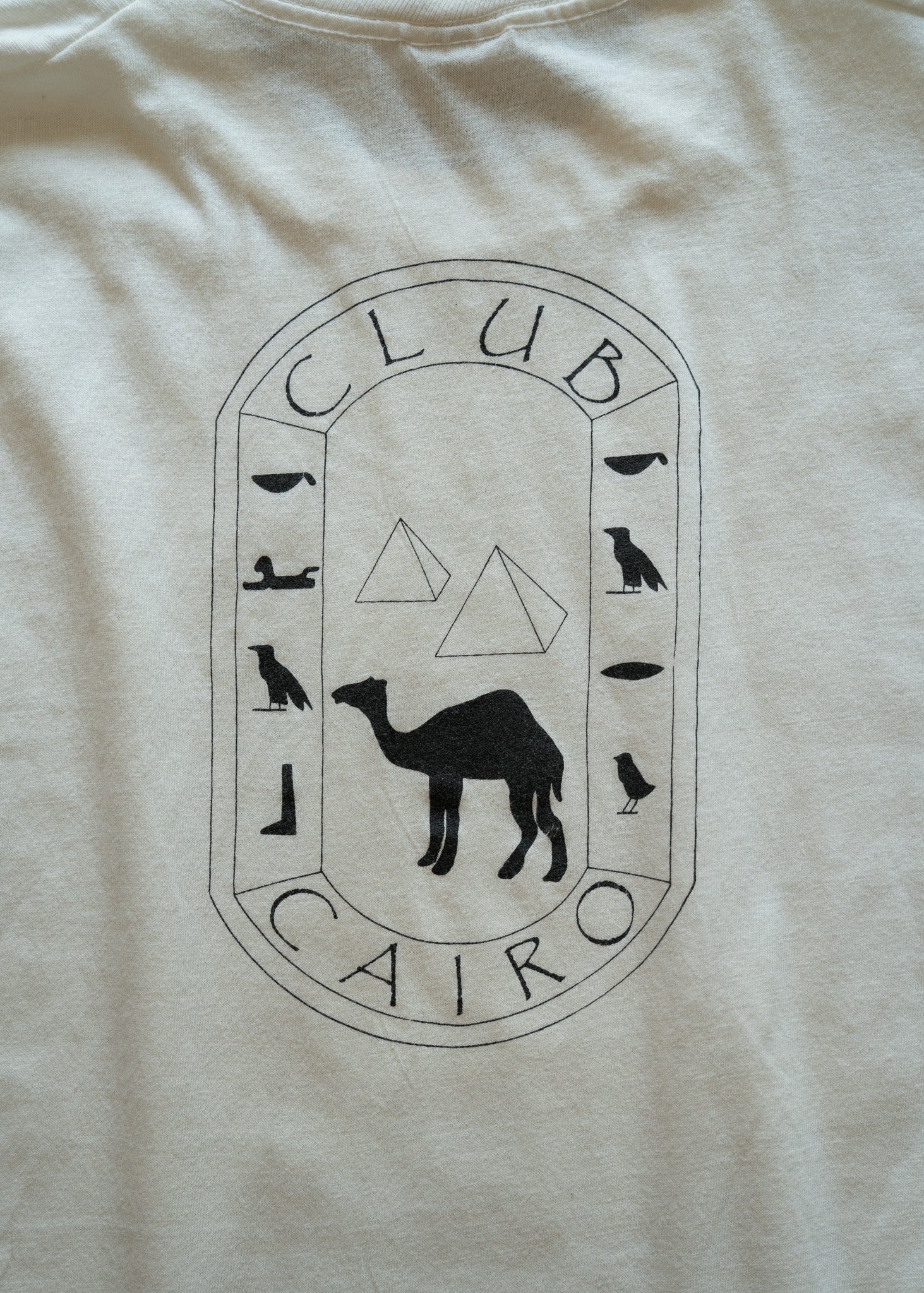 Beige t-shirt with 'Club Cairo' logo featuring a camel and Egyptian hieroglyphs.