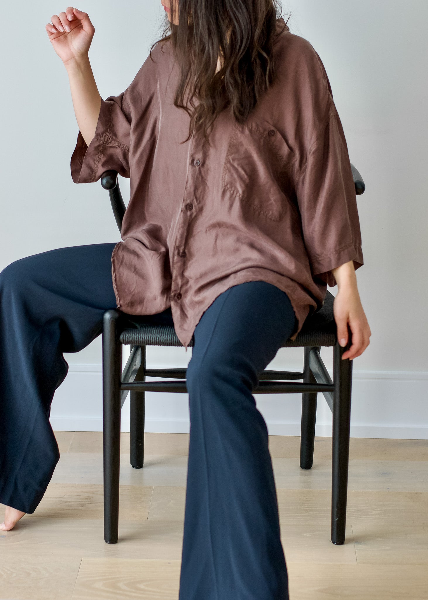 Person wearing a brown silk shirt and navy pants sitting on a chair against a plain wall.