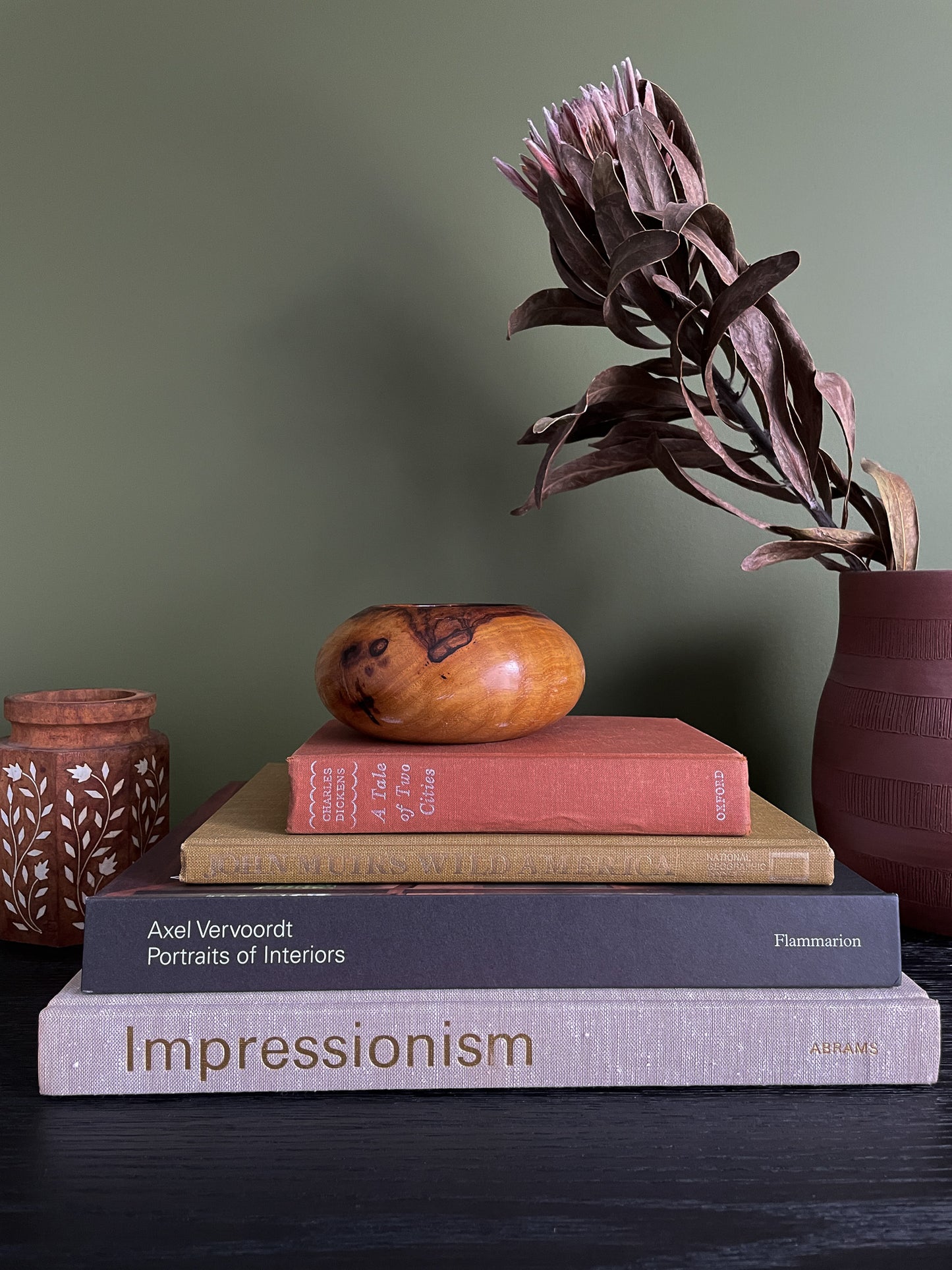 Stack of books with a decorative vintage wooden bowl and plant on a dark surface against a green wall.