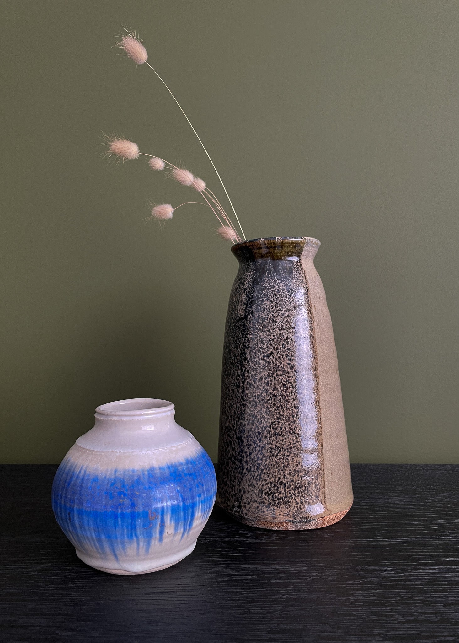 Two vintage ceramic vases with textured surfaces and dried flowers on a dark surface against a green background