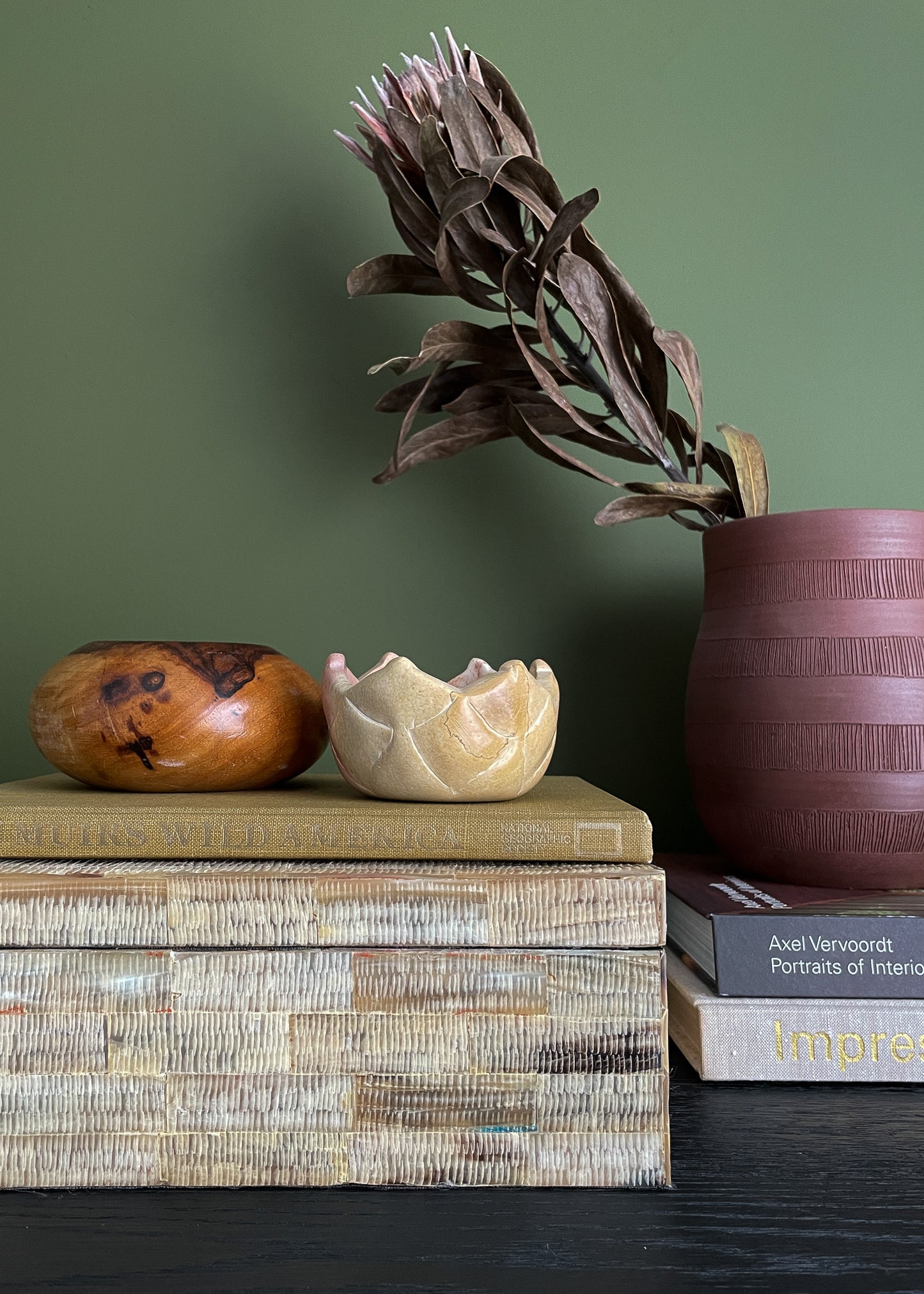 Decorative items including a wooden bowl, ceramic piece, vintage shell box and dried plant on a stack of books against a green wall.